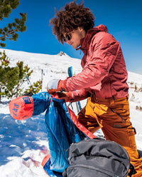 A man on a snowy mountain stuffs his ultra-light Backcountry Puffy Blanket back into its stuff sack.