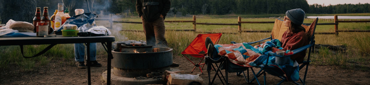 A person sits in a camp chair by the campfire, wrapped in their Geo Cool Original Puffy Blanket for warmth and comfort.