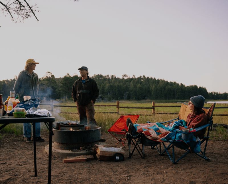 A person sits in a camp chair by the campfire, wrapped in their Geo Cool Original Puffy Blanket for warmth and comfort.