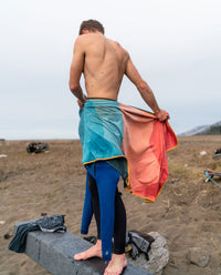 A person using a Rumpl quick-drying towel to dry off and cover up while taking off their wetsuit.